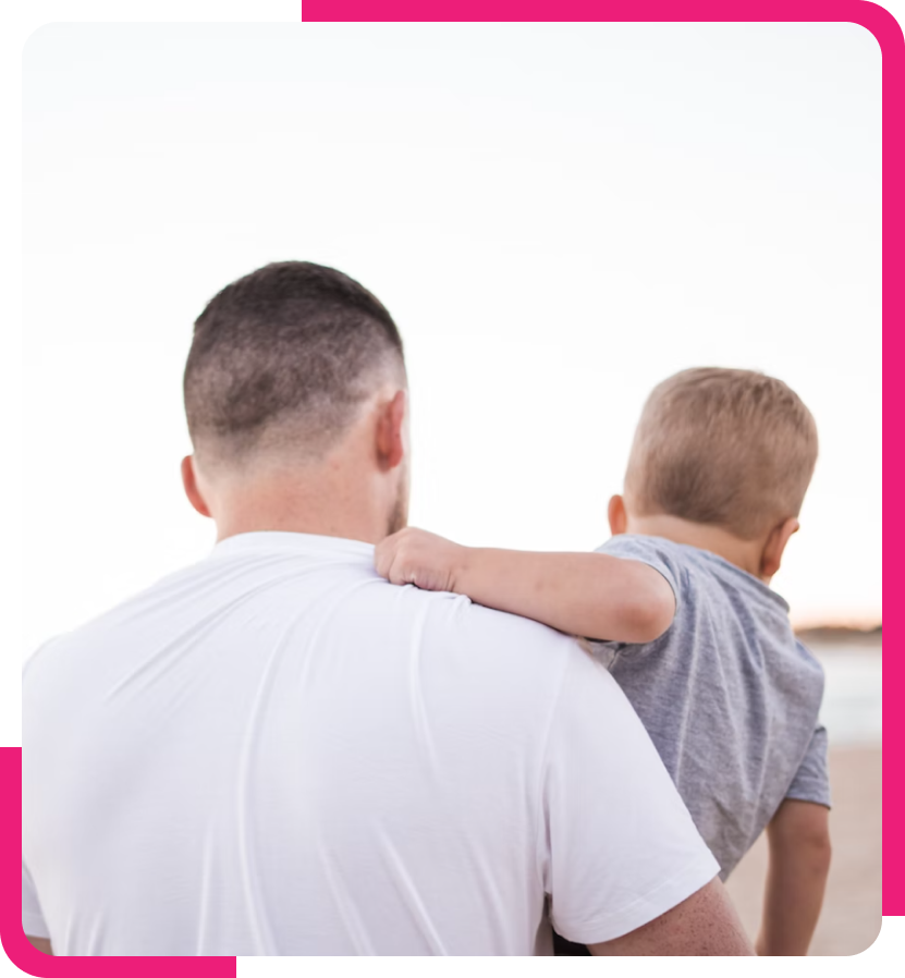 The image shows a rear view of a father carrying his young child on his back. Both are wearing casual tops; the father is in a white t-shirt, and the child in a gray one. The father's haircut is short and practical, while the child's light hair is just barely visible from this angle. They seem to be looking off into the distance, suggesting a moment of shared contemplation or observation. The background is overexposed, implying they might be outdoors. The image is framed with a pink border, giving it a warm, gentle feel.