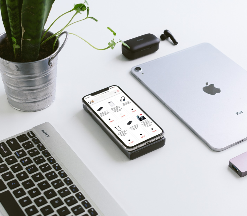 The image shows a modern, minimalist workspace setup with a smartphone displaying a shopping app, wireless earbuds in a charging case, a potted plant, and an iPad and keyboard in the background. The focus is on the smartphone in the foreground which sits next to the keyboard, suggesting a work environment that incorporates technology and greenery. The overall aesthetic is clean and organized, with white being the predominant color accented by the green plant and black tech accessories.