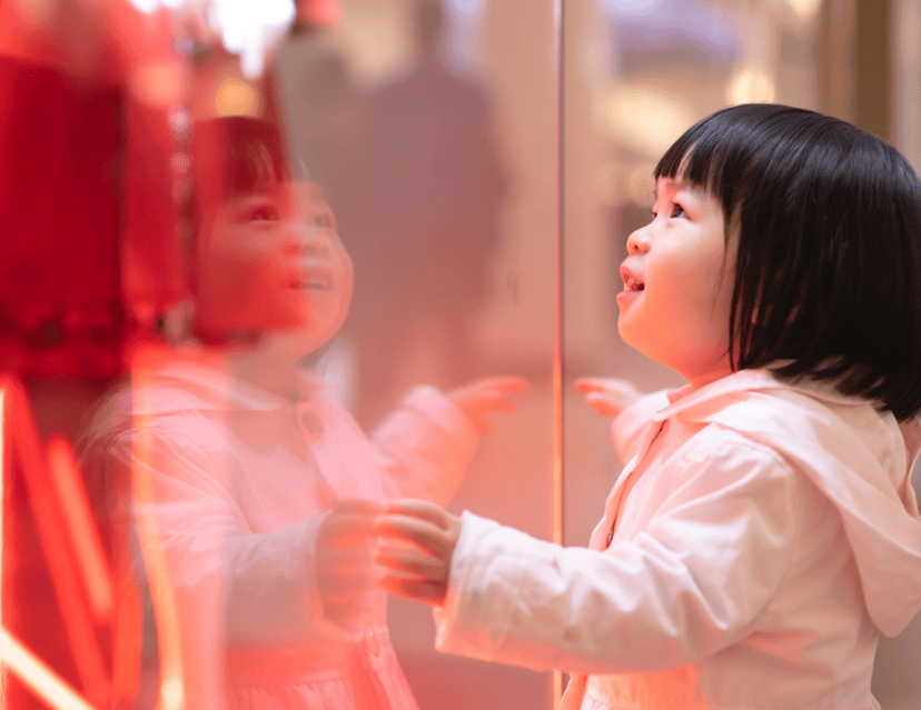 The image shows a young child with dark hair in a light jacket, looking up in wonder at something outside of view. Her reflection is visible on a shiny surface, possibly a window, which adds a dreamy quality to the image. The warm lighting casts a soft glow on her face, emphasizing her expression of innocent curiosity.