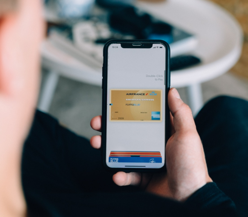 The image captures a person's hands holding a smartphone with a mobile payment screen displayed. An Air France American Express card is visible on the phone screen, indicating a transaction in progress with a prompt to double-click to pay. The setting suggests a casual environment with a blurred background that might be an indoor space.
